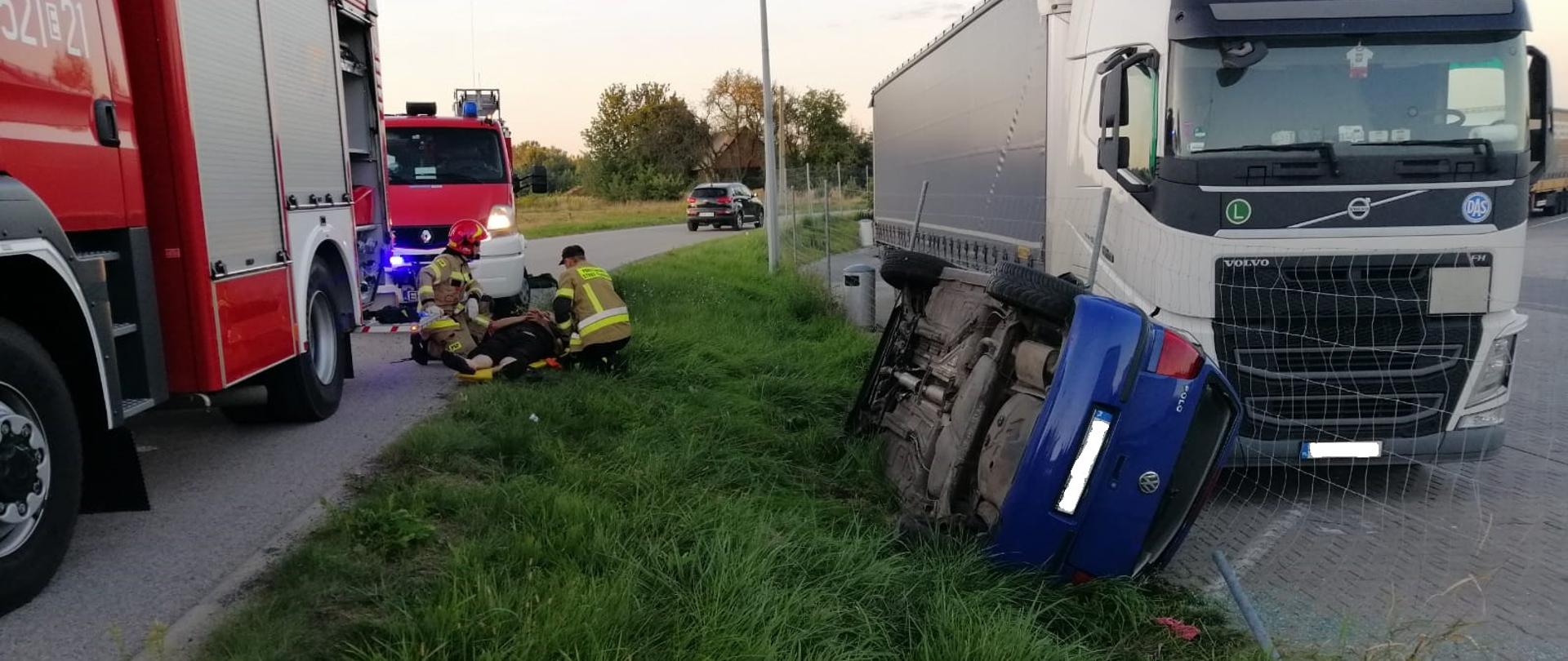 Niebieski samochód osobowy leży na prawym boku, na pasie zieleni między drogą (po lewej stronie), a parkingiem na samochody (po prawej stronie). Samochód opiera się o siatkę ogrodzenia. Za samochodem na parkingu stoi biały tir. Po lewej stronie na drodze stoją dwa samochody strażackie, pomiędzy nimi na trawie, na noszach leży poszkodowany, a obok niego jest dwóch strażaków udzielających pierwszej pomocy.