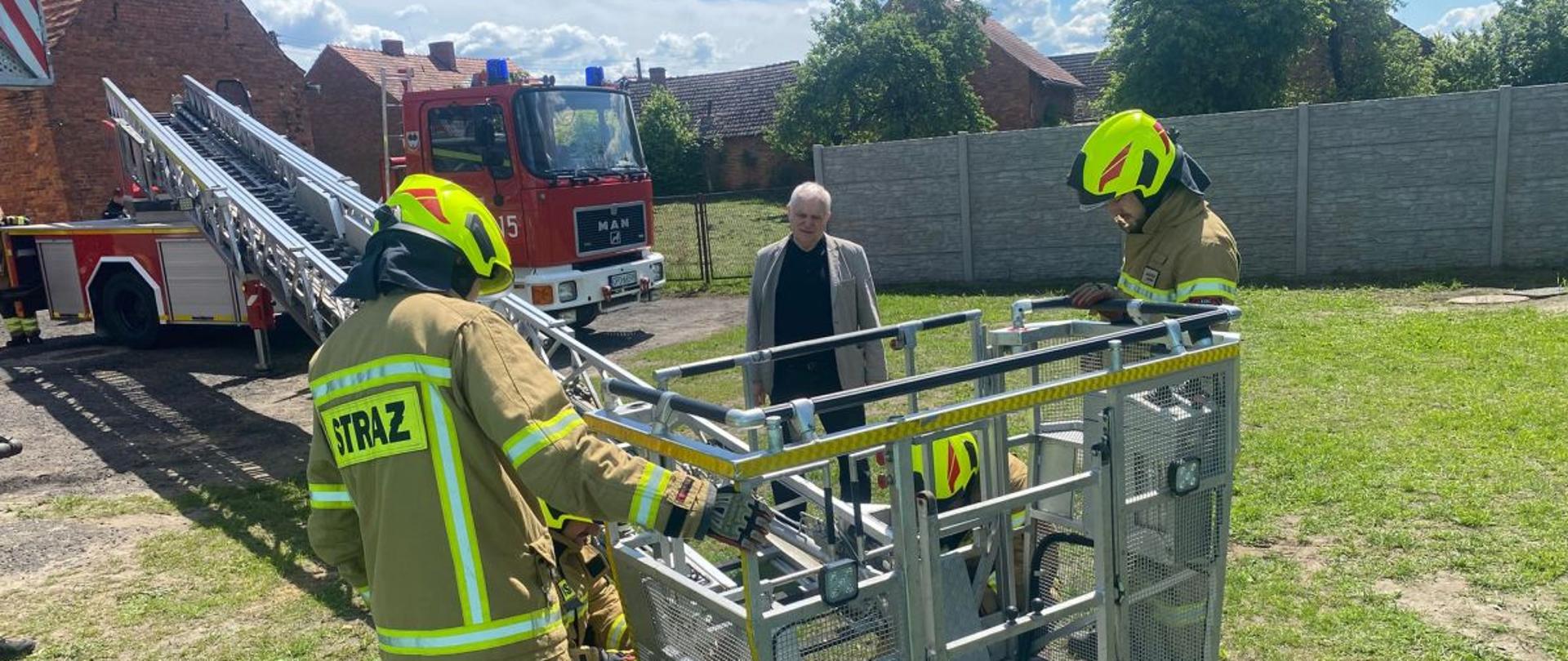 Pojazd pożarniczy typu drabina mechaniczna stoi za budynkiem. Pojazd jednostki OSP z terenu województwa opolskiego koloru czerwonego. Strażacy OSP w umundurowaniu bojowym stoją i zapoznają się z budową oraz funkcjonalnością kosza ratowniczego. Drabina rozłożona z koszem przy ziemi. W tle widoczny wykładowca i prowadzący szkolenie.