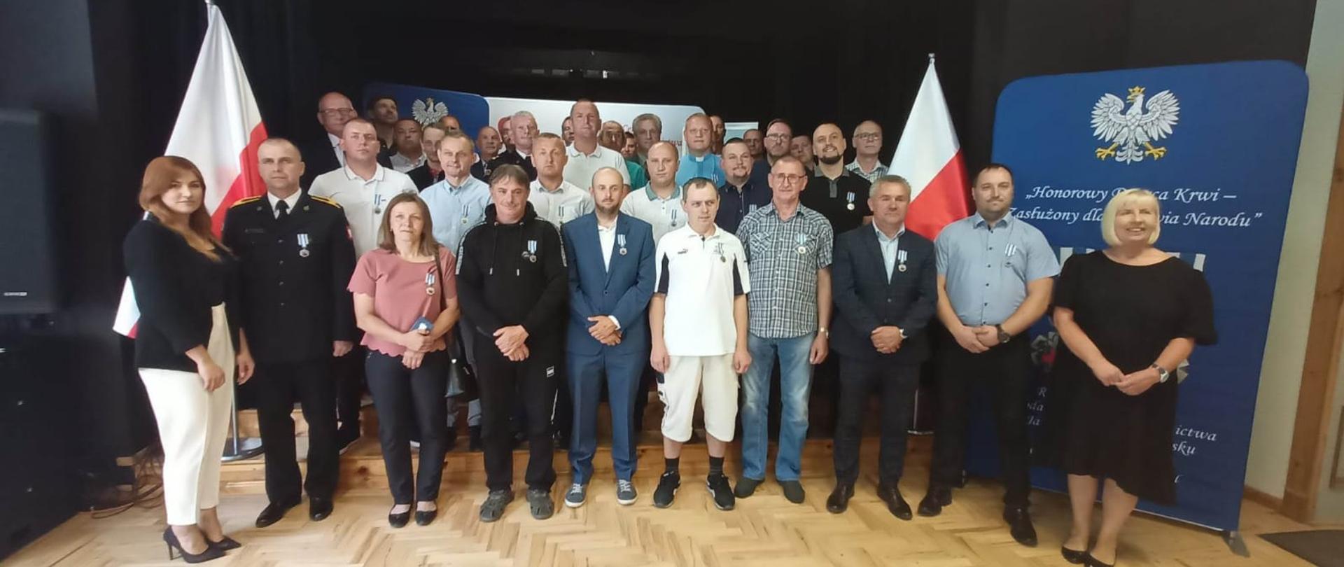 Grupka ludzi pozuje do zdjęcia. Większość z nich ma zawieszone odznaczenia po lewej stronie piersi. Po bokach postawione są biało czerwone flagi Polski.