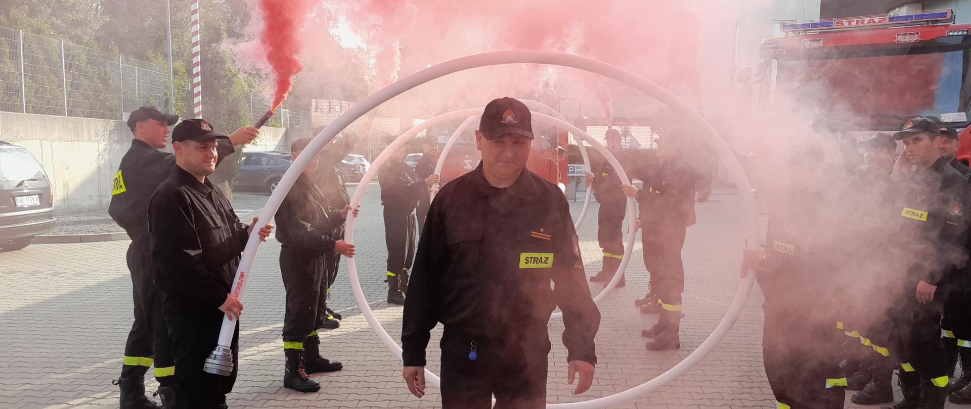 Uroczysta zmiana służby strażaka odchodzącego na zaopatrzenie emerytalne. Strażak przechodzący przez wąż strażacki zwinięty w okręgi trzymany przez strażaków, w otoczeniu czerwonego dymu z rac dymowych trzymanych przez strażaków. w tle Samochód strażacki.