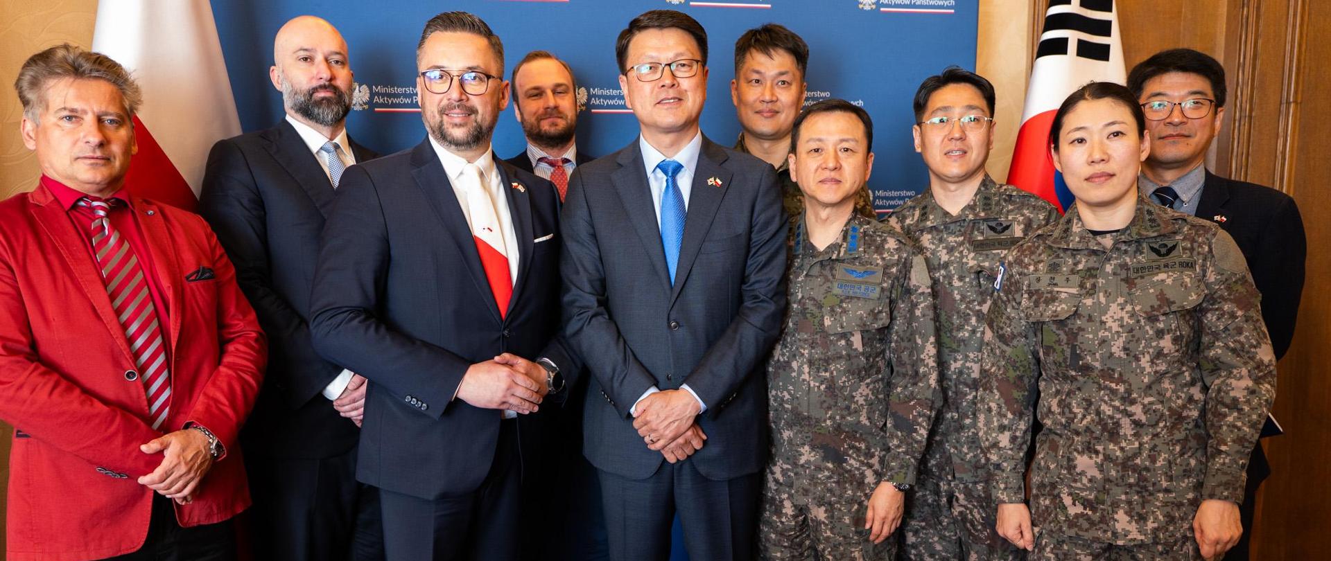 Zdjęcie grupowe członków obydwu delegacji. W tle ścianka z logotypami MAP oraz flagi Polska i Koreańska. Wiceminister Marcin Kulasek i Minister Koreański stoją w środku.
