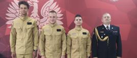 Na zdjęciu widzimy cztery osoby: trzech młodych mężczyzn w identycznych piaskowych mundurach służbowych typu taktycznego z pustymi rzepami na naszywki oraz jednego starszego oficera w tradycyjnym galowym mundurze wyjściowym Straży Pożarnej. Stoją oni na drewnianej podłodze na tle dużej czerwonej ścianki ekspozycyjnej z białym napisem „Państwowa Straż Pożarna” oraz „Komenda Powiatowa psp w Wołowie” oraz symbolem orła i godłem straży. Oficer po prawej stronie ma na piersi baretki odznaczeń oraz złoty sznur galowy.