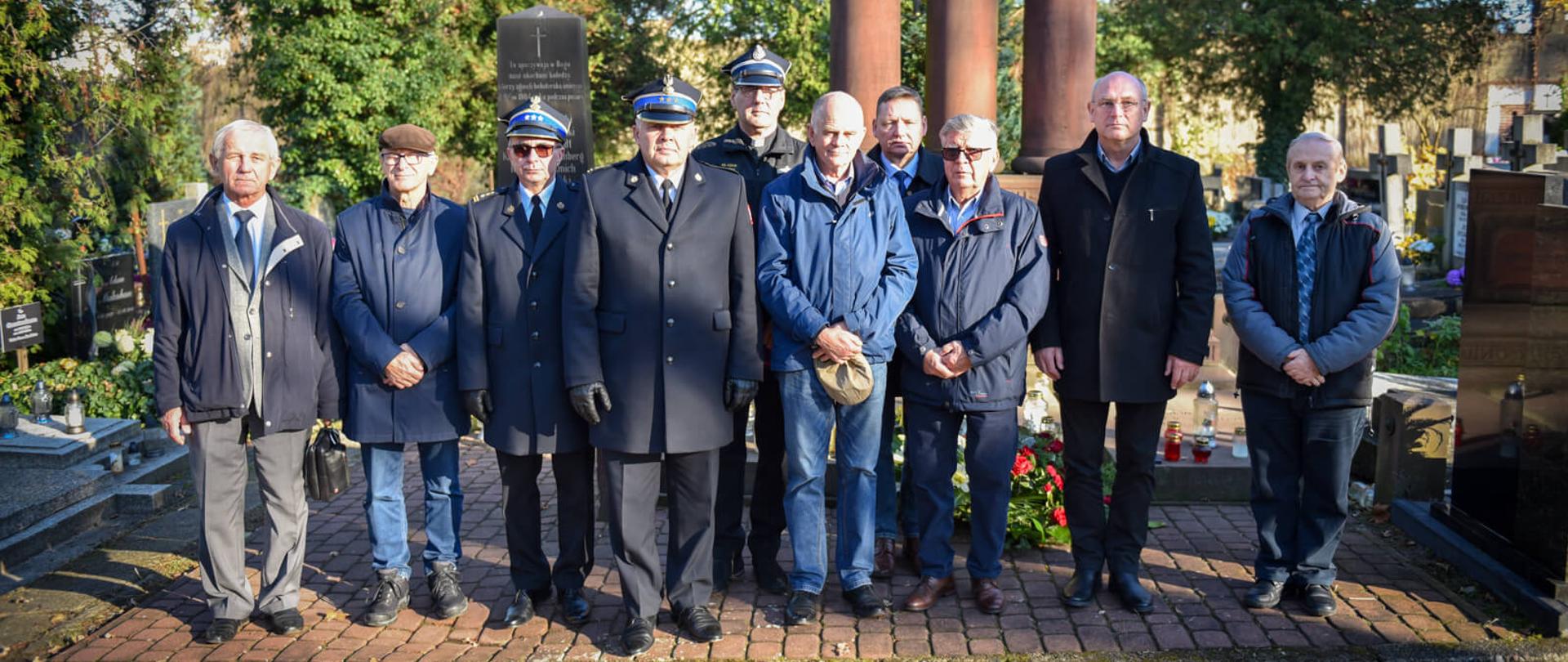 Grupę mężczyzn, w tym kilku w mundurach strażackich, pozujących razem na cmentarnej alejce przed pomnikami. Niektórzy cywile trzymają w rękach czapki
