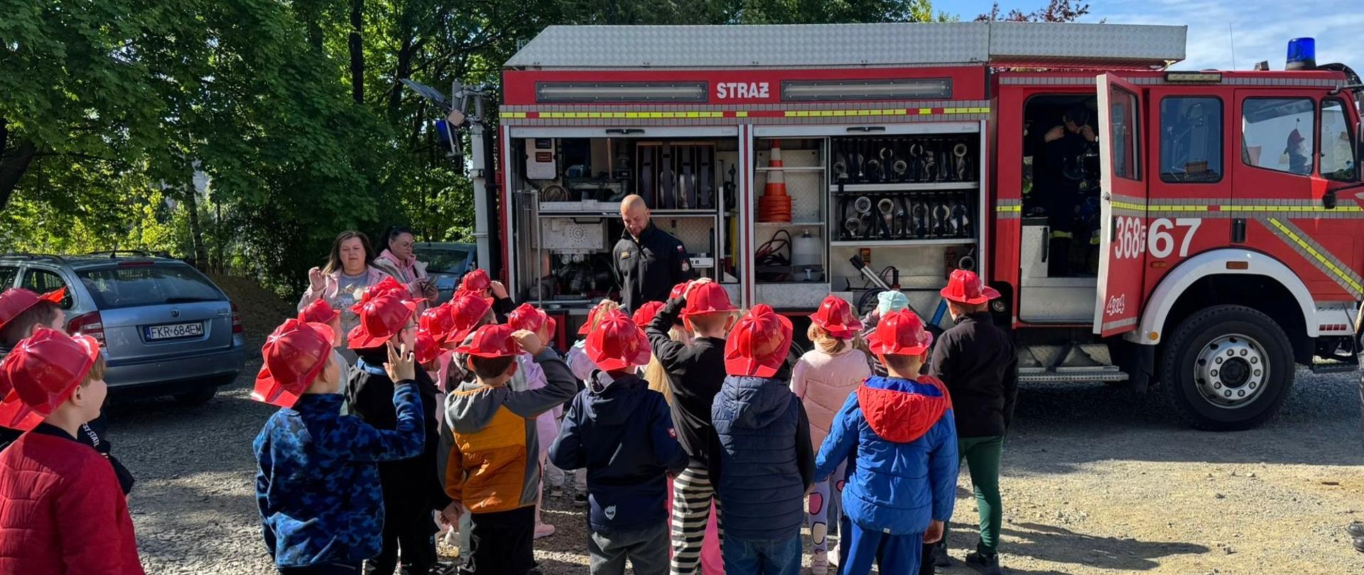 Na zdjęciu widać jak strażak pokazuje dzieciom wóz ratowniczy podczas spotkania edukacyjnego. 