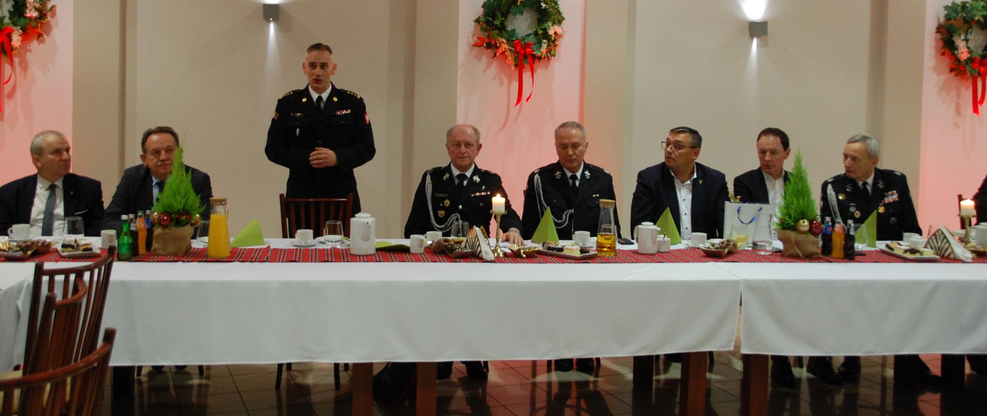 Zdjęcie zrobione w pomieszczeniu. Za stołem osiem osób, siedem siedzących oraz jedna stojąca - komendant powiatowy PSP w Lublińcu st. bryg. Janusz Bula, który przemawia. Cztery osoby w ubraniu cywilnym oraz cztery w umundurowaniu wyjściowym.