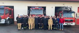 Zdjęcie przedstawia strażaków PSP podczas uroczystej zmiany służby w związku z przejściem na zaopatrzenie emerytalne Zbigniewa. W tle pojazdy pożarnicze.