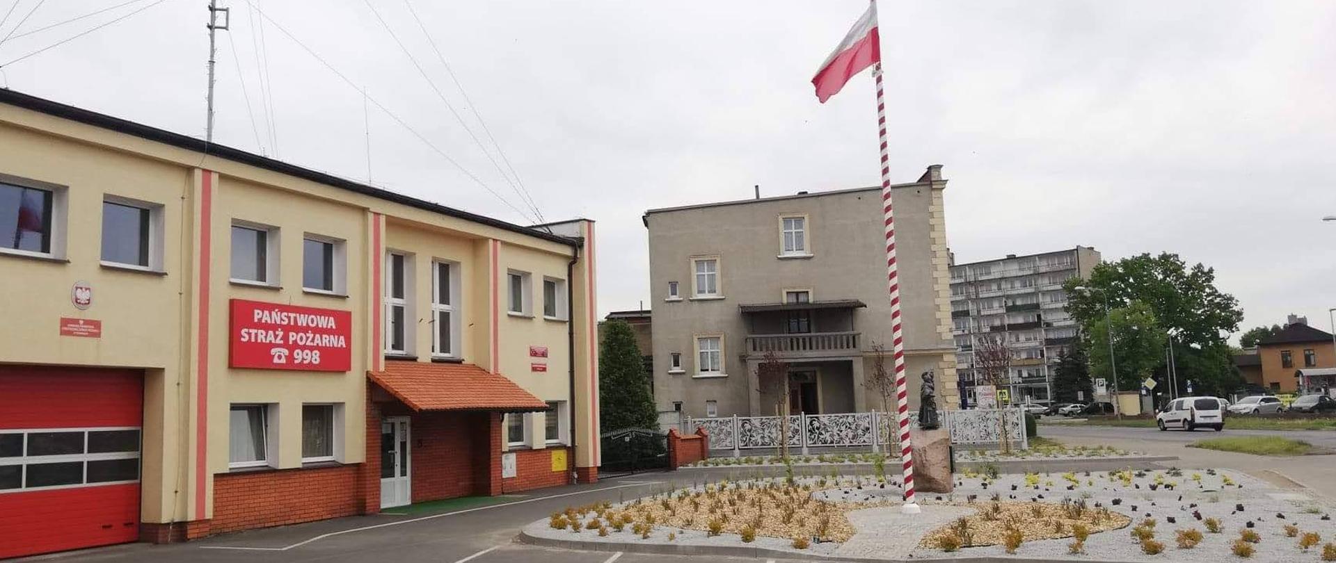 Widok skweru po rewitalizacji . Na maszcie powiewa biała flaga. Skwer zlokalizowany przed obiektem strażnicy. Duża ilość małych krzewów wypełniona kamieniem ozdobnym oraz korą. 