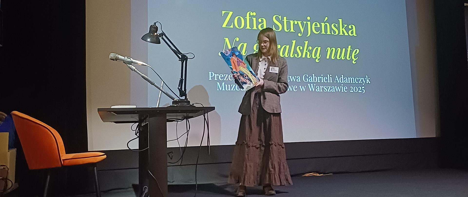 Na scenie znajduje się młoda dziewczyna, trzyma w rękach reprodukcję obrazu, z wizerunkiem górala i psa. Stoi ona na tle ogromnego ekranu z tytułem prezentacji „Zofia Stryjeńska na góralską nutę”. Na scenę prowadzą drewniane stopnie. Po lewej stronie sceny znajduje się stolik z komputerem, lampką i mikrofonem oraz pomarańczowe krzesło. 