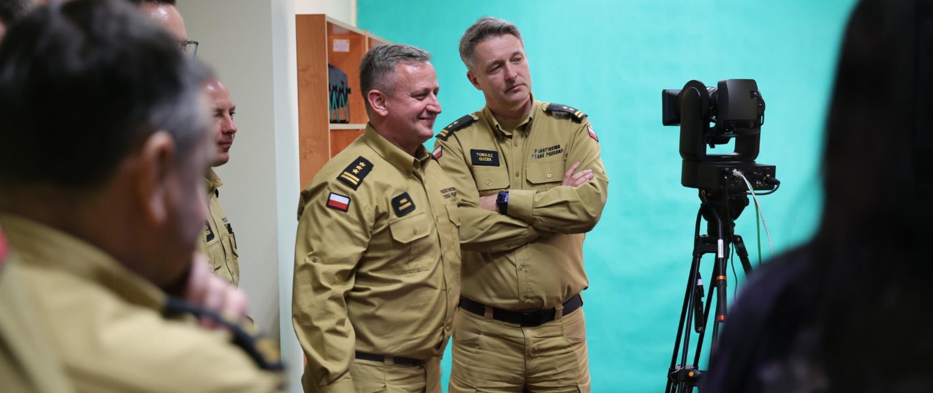 Dwóch strażaków w mundurach służbowych, w pozycji stojącej stoi przed kamerą i sa zwróceniu głowami ku siebie. Wokół kilku innych strazaków. Rzecz dzieje się w studiu tv.
