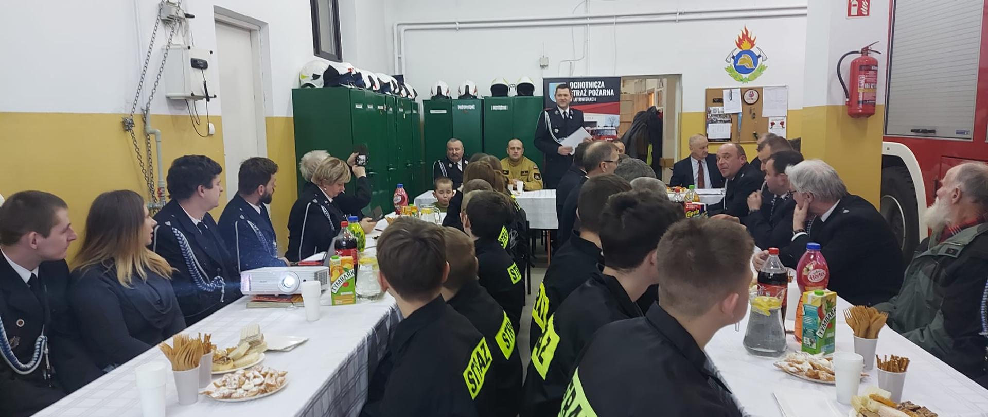 Na zdjęciu widać garaż jednostki OSP Lutowiska w którym ustawione są stoły i przy których siedzi komendant powiatowy PSP w Ustrzykach Dolnych w ubraniu koszarowym piaskowym oraz druhowie i druhny jednostki OSP Lutowiska. Stołów są dwa rzędy a na końcu jeden stół ustawiony jest prostopadle do nich. Widać także szafki koloru zielonego oraz kawałek samochodu GCBA Scania P400. 