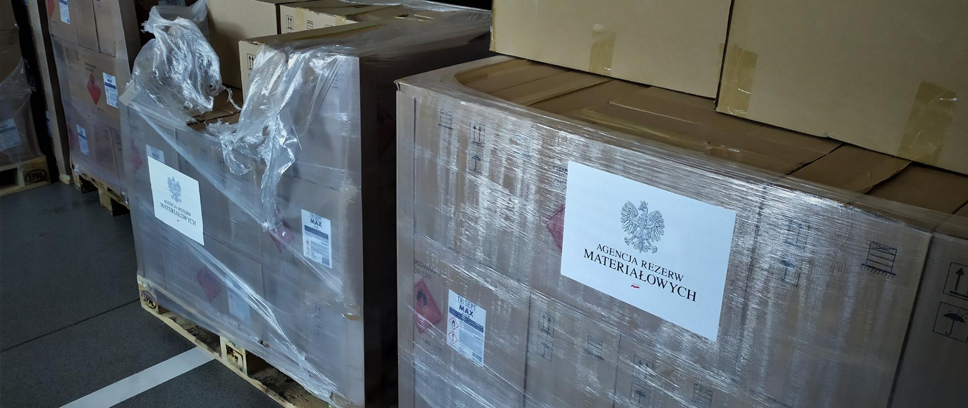 Na zdjęciu środki ochrony indywidualnej zapakowane w kartony na palecie transportowej. Na jednym z nich napis agencja rezerw materiałowych. Zdjęcie wykonane w garażu Jednostki Ratowniczo-Gaśniczej w Lubaczowie.