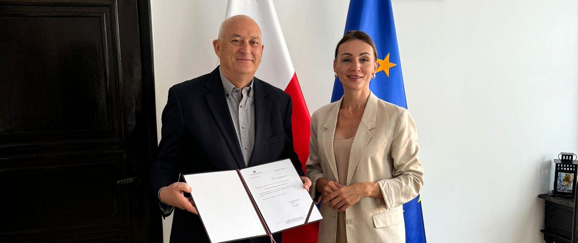 wręczenie powołania na konsultanta wojewódzkiego. Mężczyzna trzyma w dłoniach akt nominacji obok stoi kobieta, za nimi flagi polska i unijna