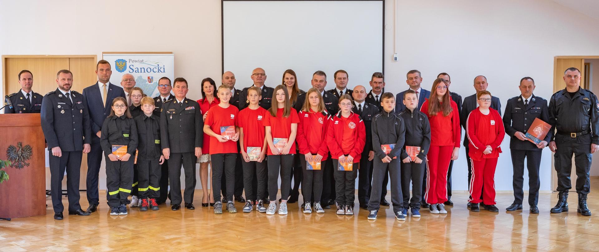 Zdjęcie wykonane wewnątrz pomieszczenia. Zdjęcie przedstawia wszystkich uczestników uroczystości - zdjęcie grupowe. 