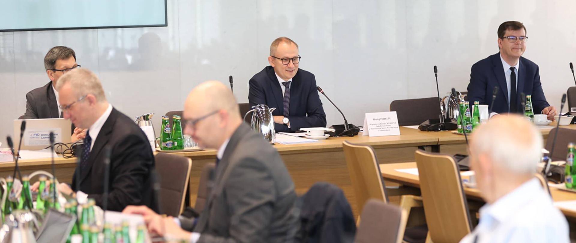 Wiceminister Błażej Poboży siedzący na prezydium obradującej Komisji Wspólnej Rządu i Mniejszości Narodowych i Etnicznych.