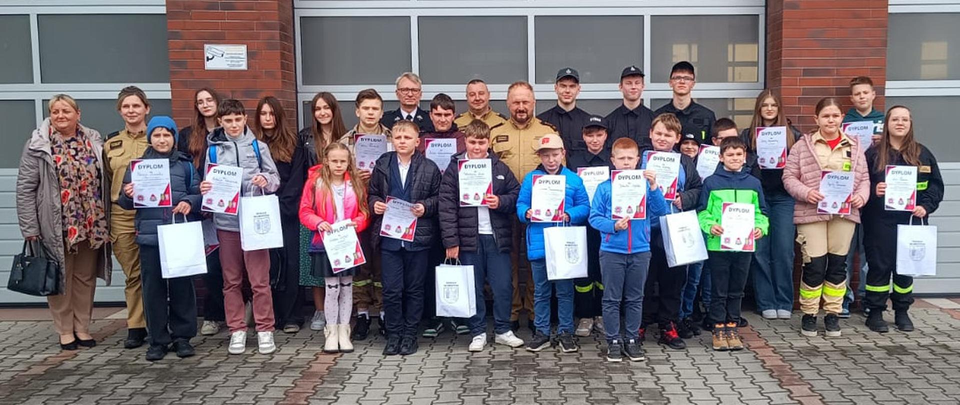 Duża grupa dzieci i młodzieży – uczestników turnieju pożarniczego – stoi przed bramą garażową strażnicy w Głubczycach. Trzymają w rękach białe dyplomy i papierowe torby z nagrodami. W tylnym rzędzie stoją strażacy w mundurach, w tym Komendant Powiatowy Państwowej Straży Pożarnej w Głubczycach oraz opiekunowie.