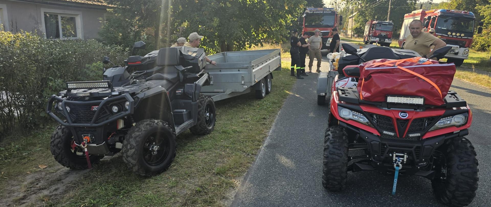 Na pierwszym zdjęciu widać dwa quady strażackie, obok nich strażacy. Za nimi są trzy wozy strażackie. Wszyscy znajdują się w pobliżu drogi, gdzie są domy mieszkalne.