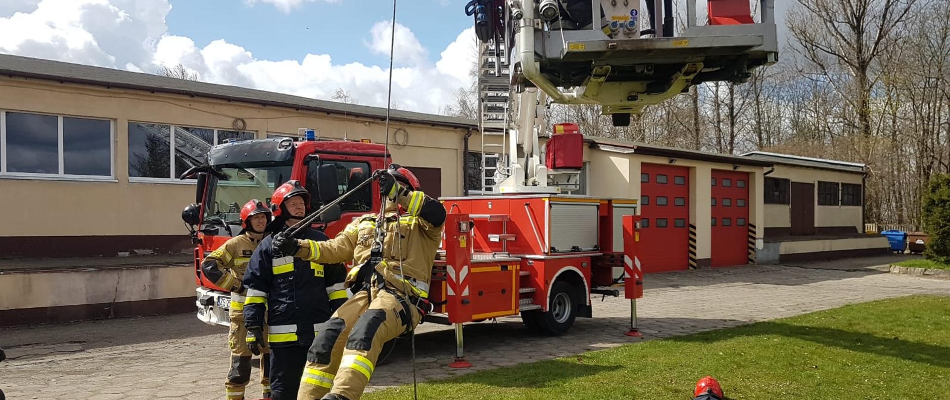 Zdjęcie przedstawia samochód specjalny z drabina hydrauliczną rozstawiona oraz strażaków ćwiczących z linami ratowniczymi.
