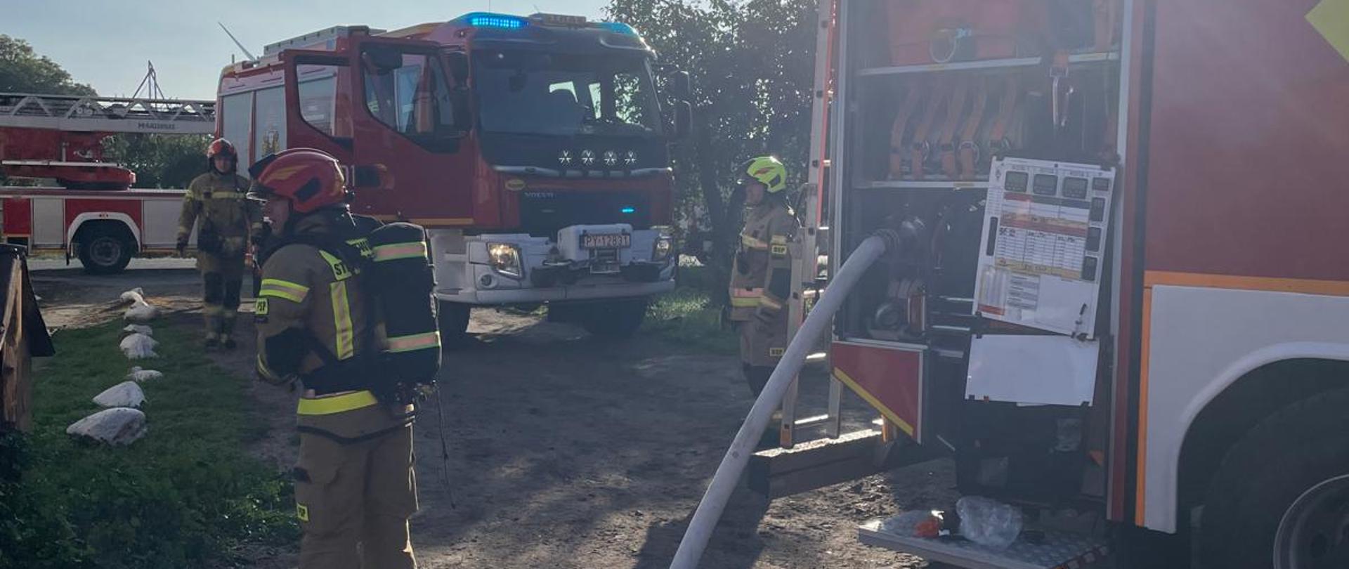 Zdjęcie przedstawia strażaków podczas pożaru w budynku wielorodzinnym.
W tle samochody strażackie.
Zdjęcie przedstawia butle propan-butan, które zostały wyniesione z budynku.
W tle tereny zielone.
