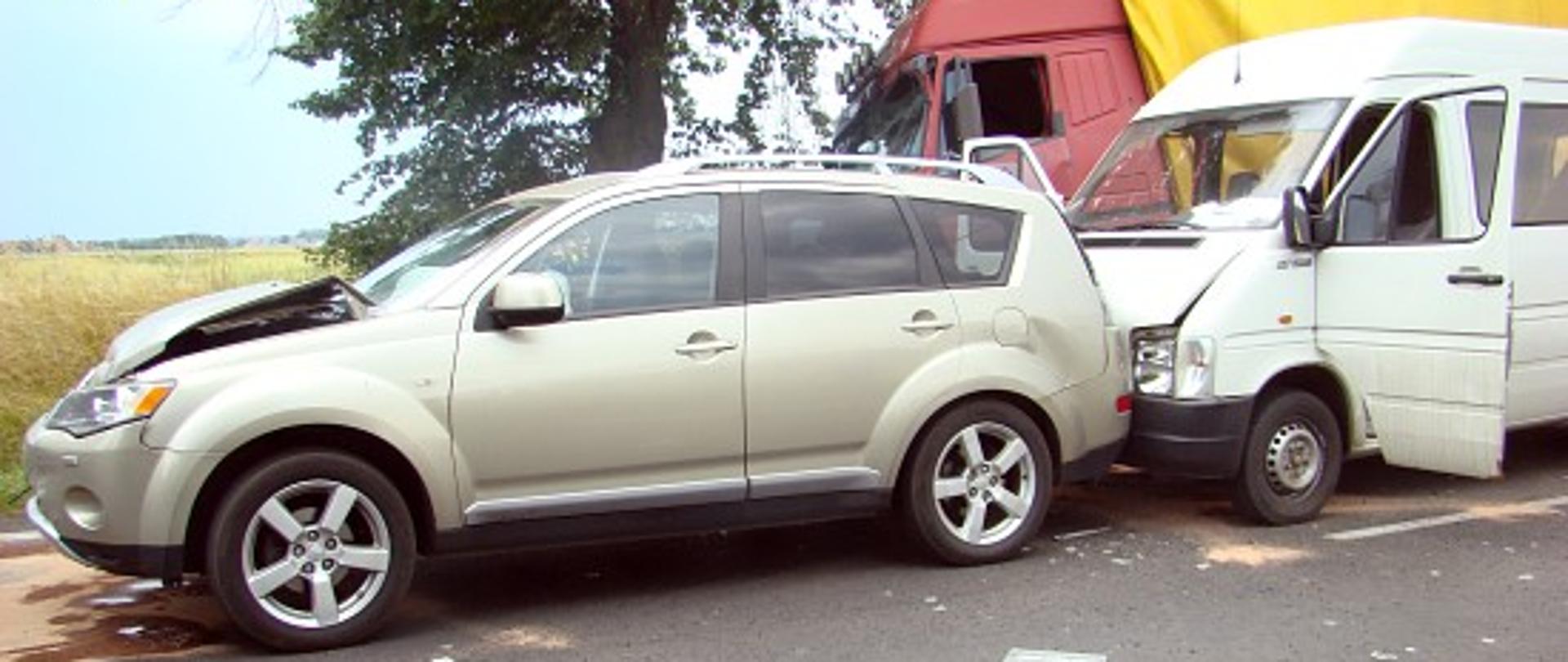 Widoczne dwa uszkodzone pojazdy osobowe stojące na jezdni. W tle lekko w rowie pojazd ciężarowy.