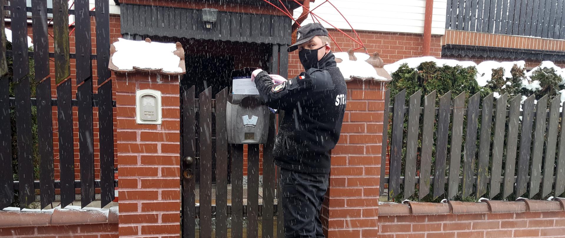 Na zdjęciu widoczna jest ulica i dom. Przy furtce stoi strażak w ubraniu koszarowym i maseczce ochronnej, który wrzuca do skrzynki na listy ulotkę dotyczącą profilaktyki oraz szczepień przeciw COVID-19. 