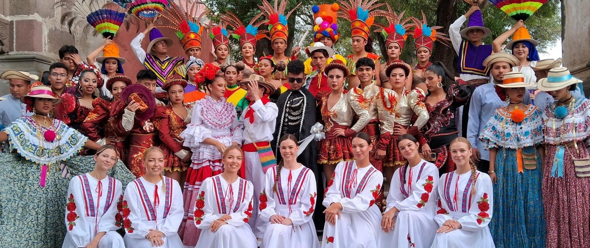 En la foto se ve a un numeroso grupo de personas con coloridos trajes tradicionales, posando para una foto conjunta al aire libre, bajo grandes árboles y junto a una arquitectura de piedra.