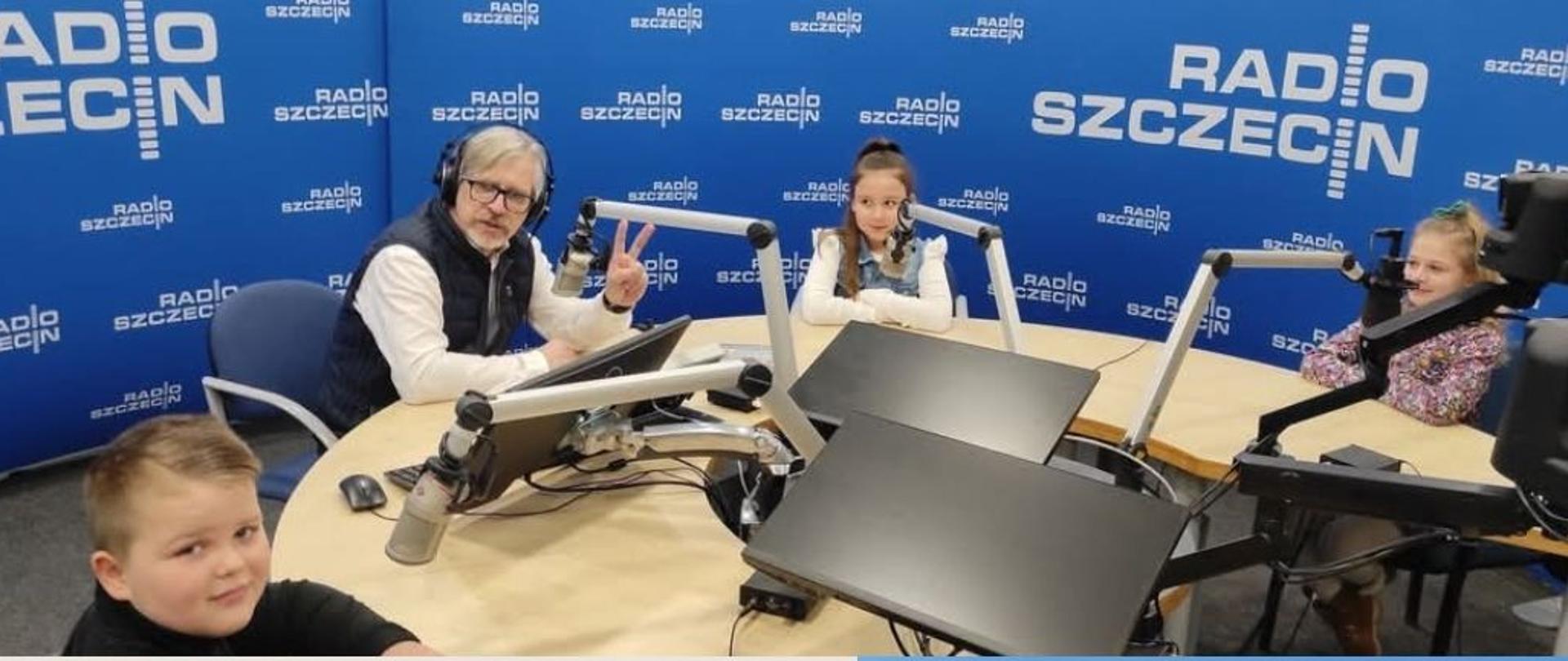 "Klub Muzycznych Odkrywców" - uczniowie PSM I st. i OSM I st. w Stargardzie w Radiu Szczecin. Plakat przedstawia collage zdjęć ze studia Radia Szczecin. Prowadzący audycję pan Paweł Osuchowski oraz uczniowie siedzą za okrągłym stołem w studiu Radia Szczecin, przed każdym z nich jest mikrofon. W tyle widać niebieską ścianę studia z białym logo Radia Szczecin.