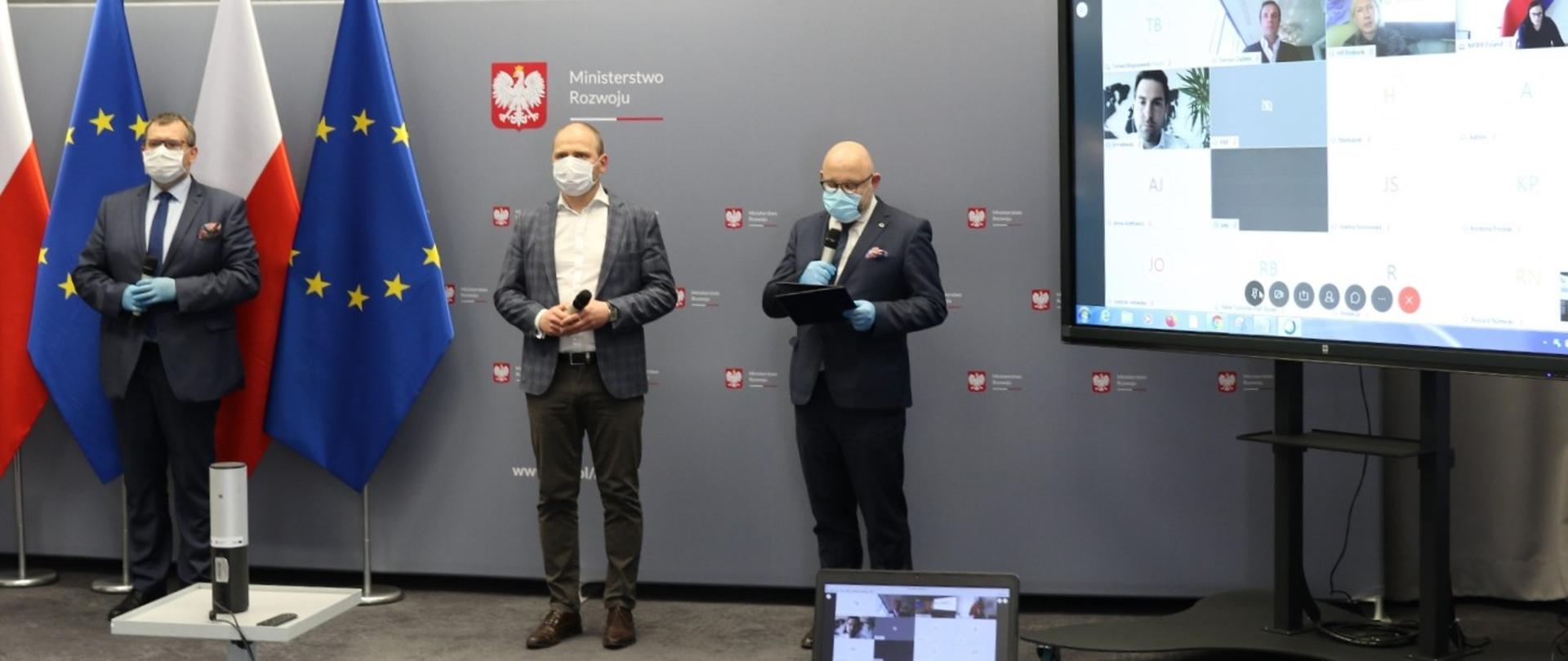 Konferencja prasowa. Po lewej stronie stoi wiceprezes ARP Kolczyński, w środku wiceminister Mazur, a z prawej strony dyrektor Sokoliński z ARP. Panowie mają na twarzach maseczki ochronne. Skrajnie po prawej stronie widać ekran z uczestnikami telekonferencji.