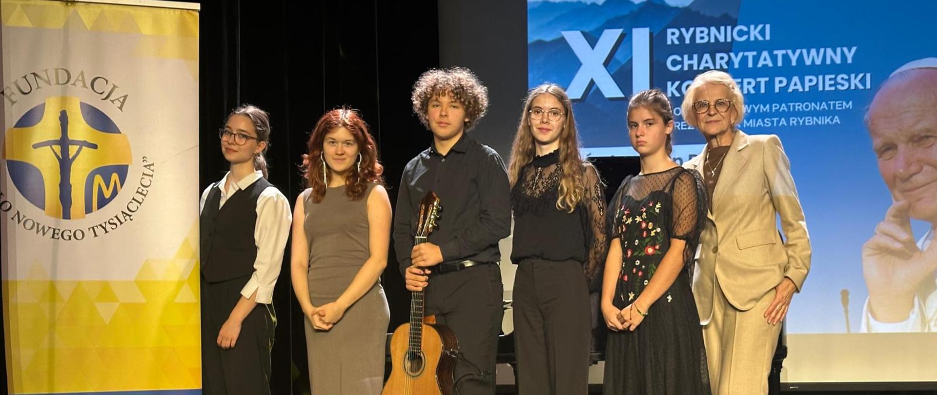 Kolorowe zdjęcie na którym stoi w jednym rzędzie 6 osób tj. młodzi wykonawcy i pani wicedyrektor szkoły. Wszyscy ubrani w eleganckie stroje, uśmiechnięci, jeden uczeń trzyma w rękach gitarę. Grupa stoi obok baneru fundacji papieskiej na tle grafiki XI Rybnickiego Charytatywnego Koncertu Papieskiego (niebieskie tło z zdjęciem Papieża Jana Pawła II). 