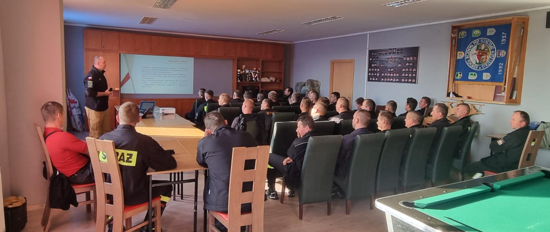 Na zdjęciu znajdują się strażacy siedzący na krzesłach, patrzą na ekran na którym wyświetlana jest prezentacja