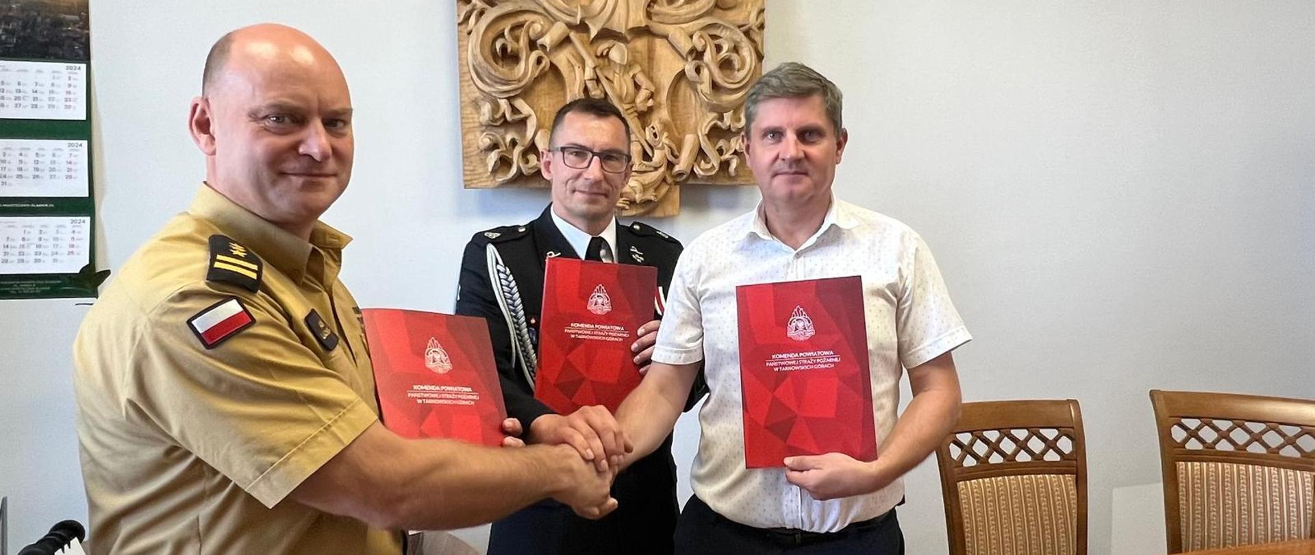 Trzech mężczyzn stoi. W rękach mają czerwone teczki, ściskają sobie dłonie.