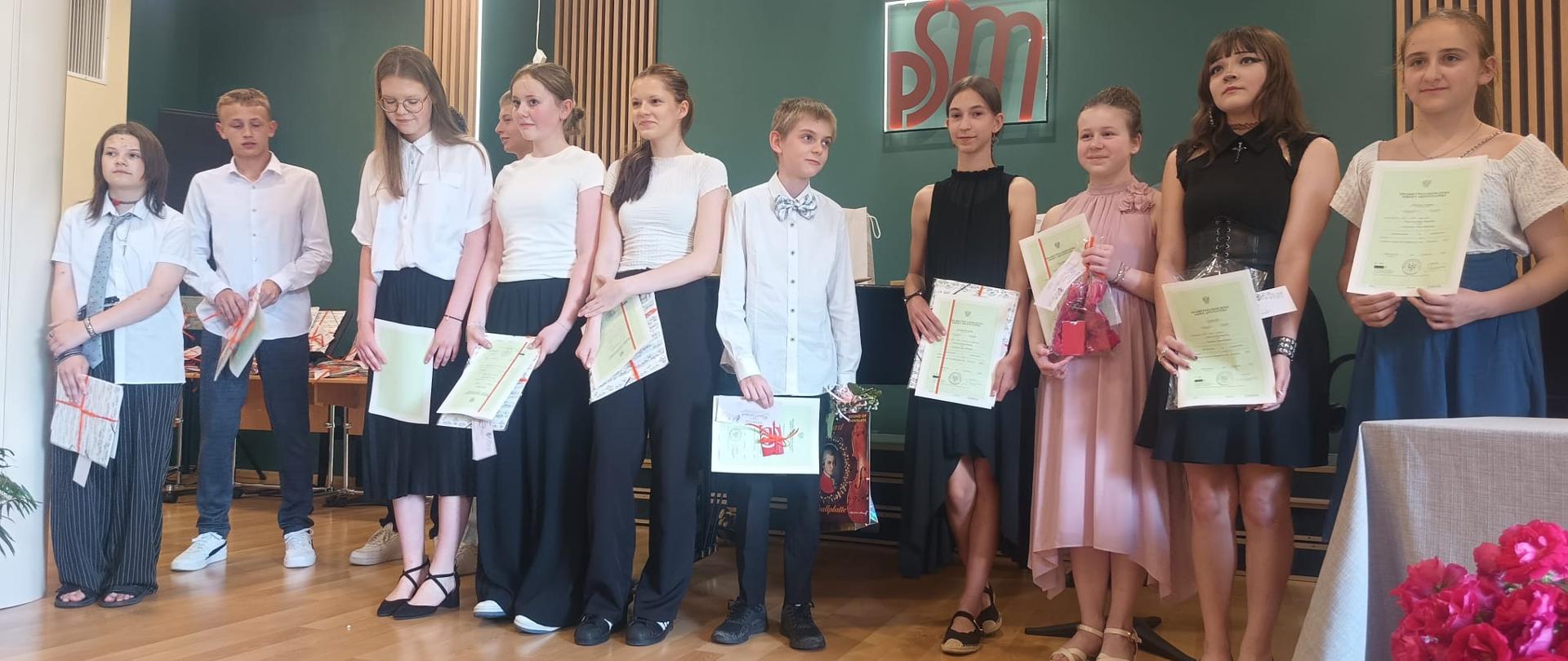 Pani dyrektor oraz nauczyciele rozdają nagrody dla uczniów z najlepszymi wynikami w nauce. Uroczystość odbywa się na sali koncertowej szkole muzycznej. Na sali są rodzice oraz uczniowie i nauczyciele.