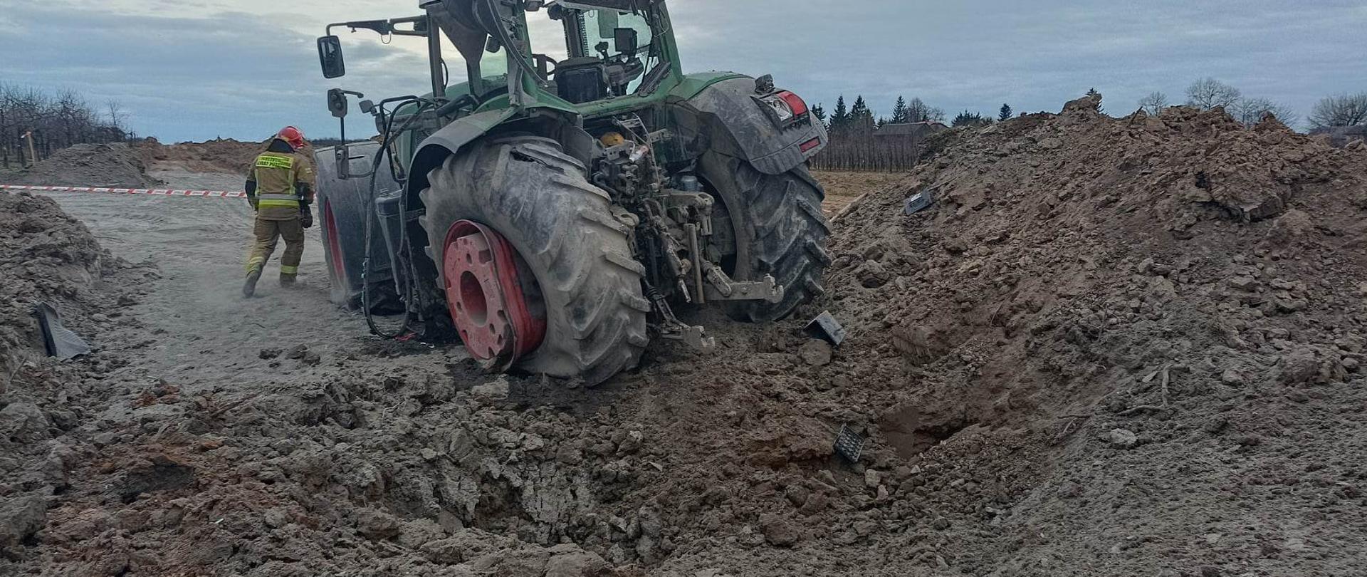 Zdjęcie przedstawia lej po eksplozji niewiadomego przedmiotu, ciągnik rolniczy, funkcjonariusz PSP 