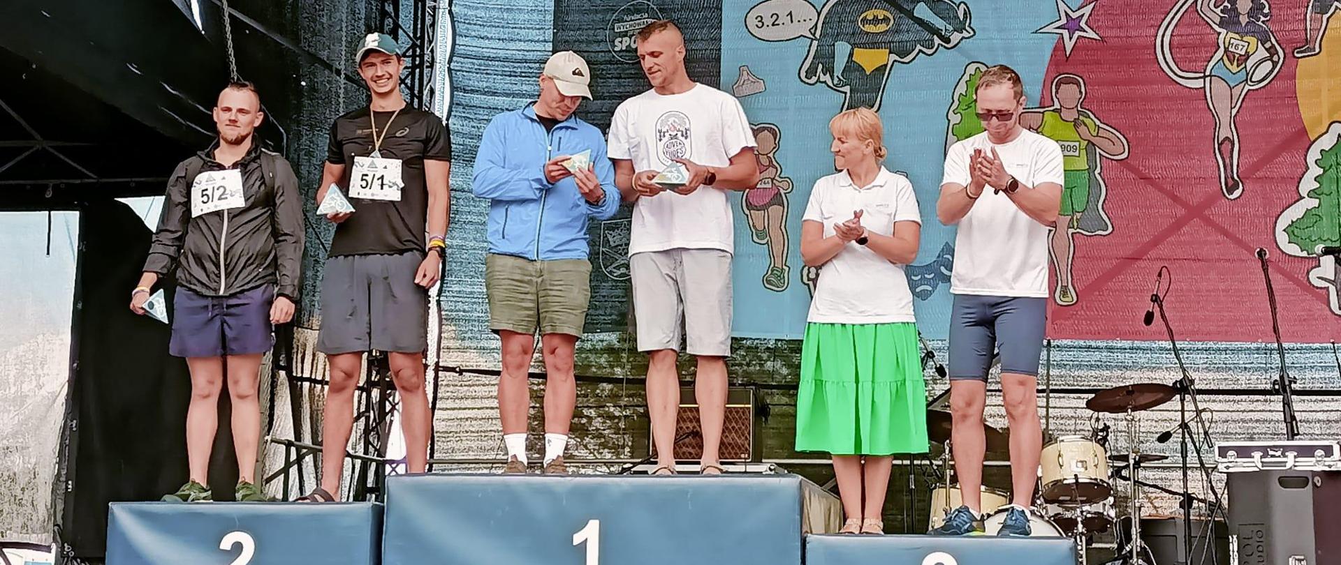 Zdjęcie przedstawia podium na którym widoczne są osoby które odpowiednio zajęły pierwsze drugie i trzecie miejsce 