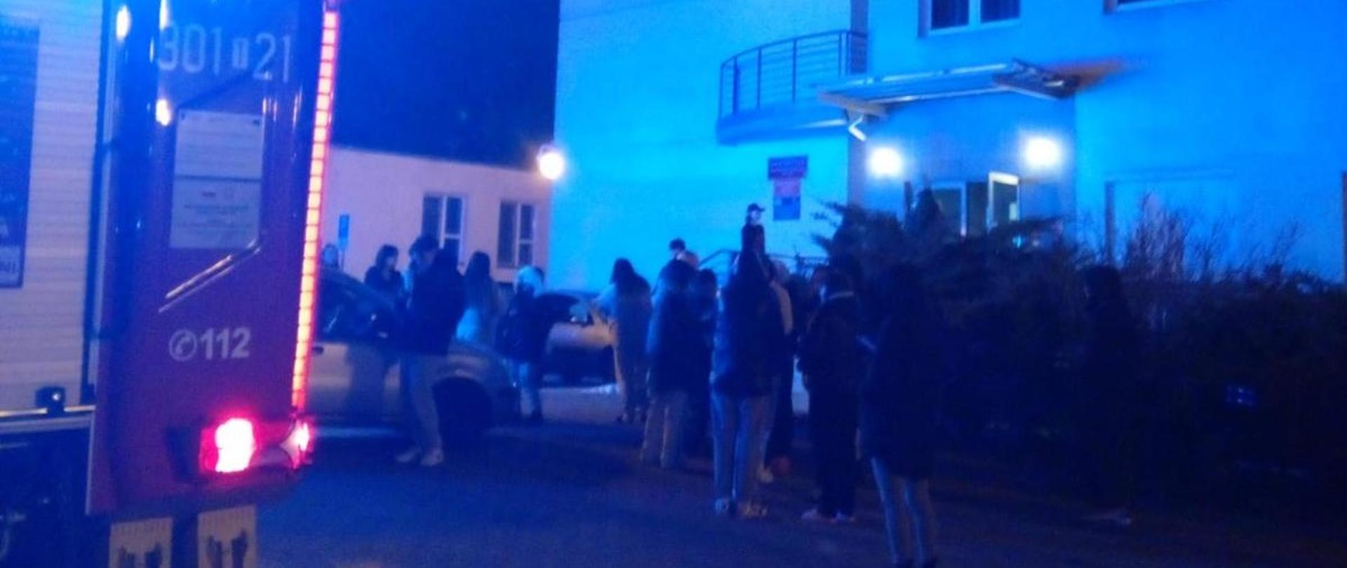 Po prawej stronie widać budynek zamieszkania zbiorowego, w którym doszło do pożaru. Przed obiektem stoją mieszkańcy, a z lewej strony widać fragment samochodu pożarniczego.