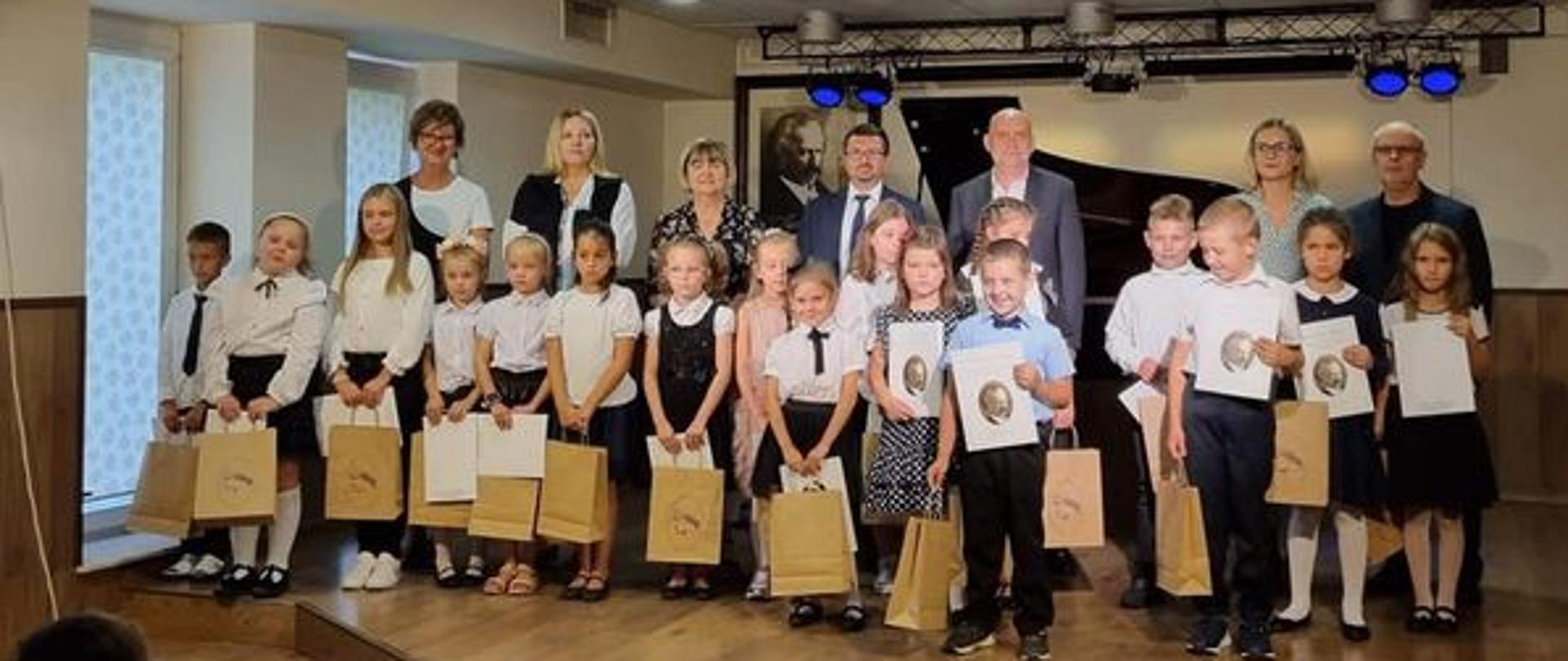 Fotografia przedstawia nowo przyjętych uczniów cyklu 6-letniego i nauczycieli. Wszyscy stoją na scenie auli szkoły. Z prawej strony widoczne 2 zasłonięte okna, za nauczycielami widać otwartą klapę od fortepianu, z tyłu na ścianie 4 niebieskie reflektory. Podłoga i boki w kolorze drewna. Na suficie 9 okrągłych lamp.