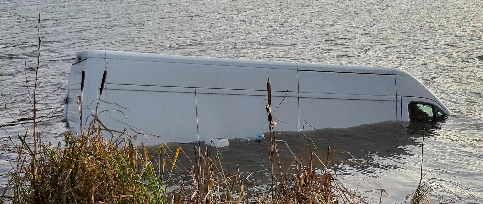 Samochód typu bus znajduj się prawie pod sam dach w wodzie przy brzegu. 