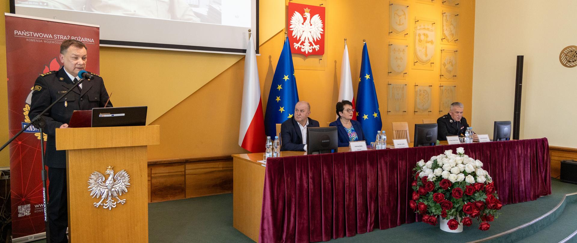 Opolski komendant wojewódzki przemawia przy mównicy. Po jego lewej stronie przy stole zasiadają goście.