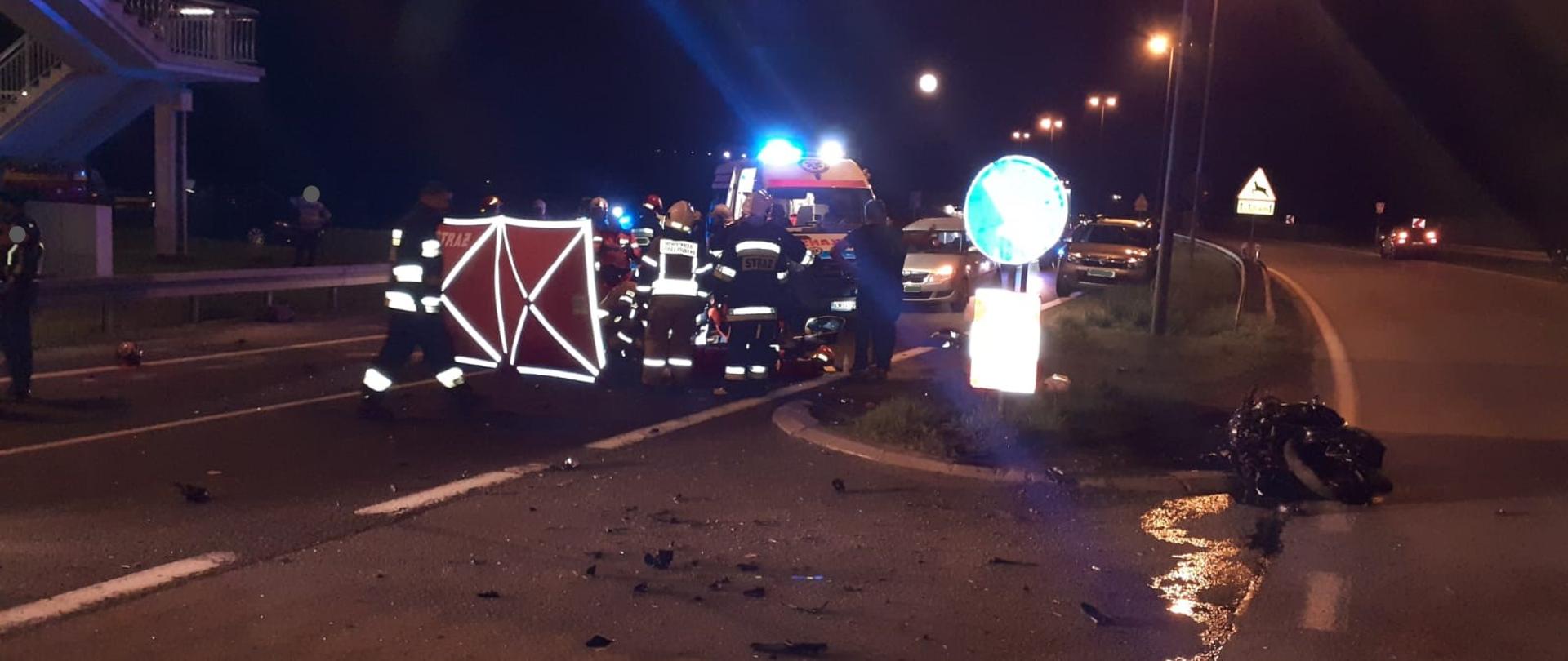 Zdjęcie wykonane w porze nocnej przedstawia drogę krajową nr 7 mającą po dwa pasy jezdni w każdym kierunku. Na skraju pasa widzimy leżący rozbity motocykl. Z lewej strony grupa strażaków, ratowników medycznych udzielających pomocy poszkodowanej w zderzeniu osobie, która znajduje się za parawanem. W tle stoi karetka pogotowia oraz pojazdy, które zatrzymały się z powodu braku przejazdu. W górnej części zdjęcia częściowo widoczna jest kładka dla pieszych w celu bezpiecznego przejścia nad droga krajową.
