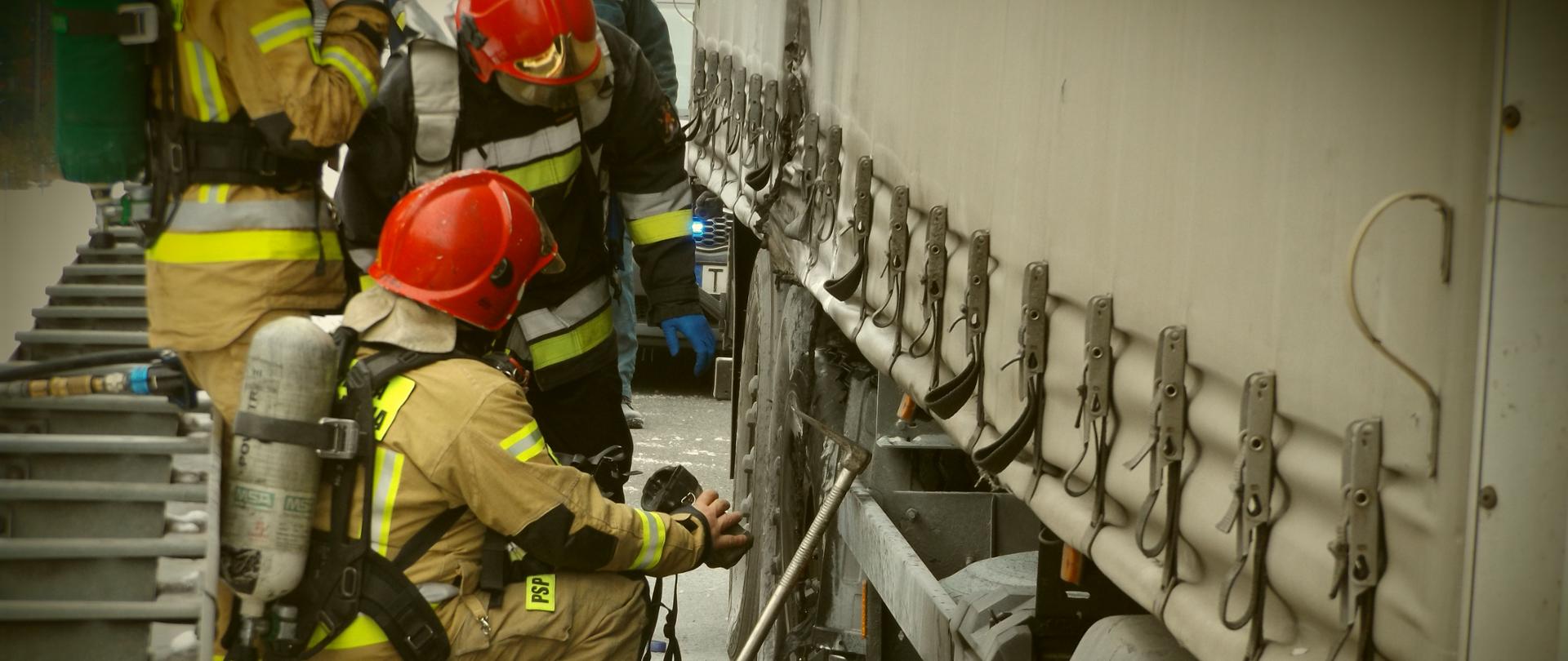 Zdjęcie przedstawia strażaków przy naczepie samochodu ciężarowego którzy za pomocą kamery termowizyjnej sprawdzają temperaturę uszkodzonej opony, potwierdzając tym samym jej całkowite ugaszenie.