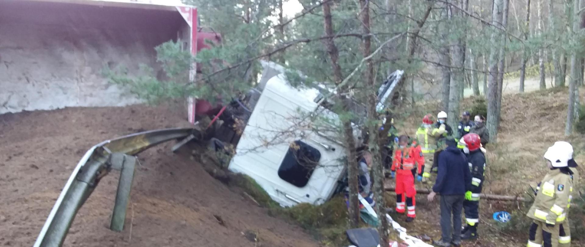 Wywrócona na bok naczepa samochodu ciężarowego, na całej szerokości jezdni rozsypany piasek. Widoczny dach kabiny pojazdu, wokół kabiny zgromadzeni ratownicy i strażacy. 
