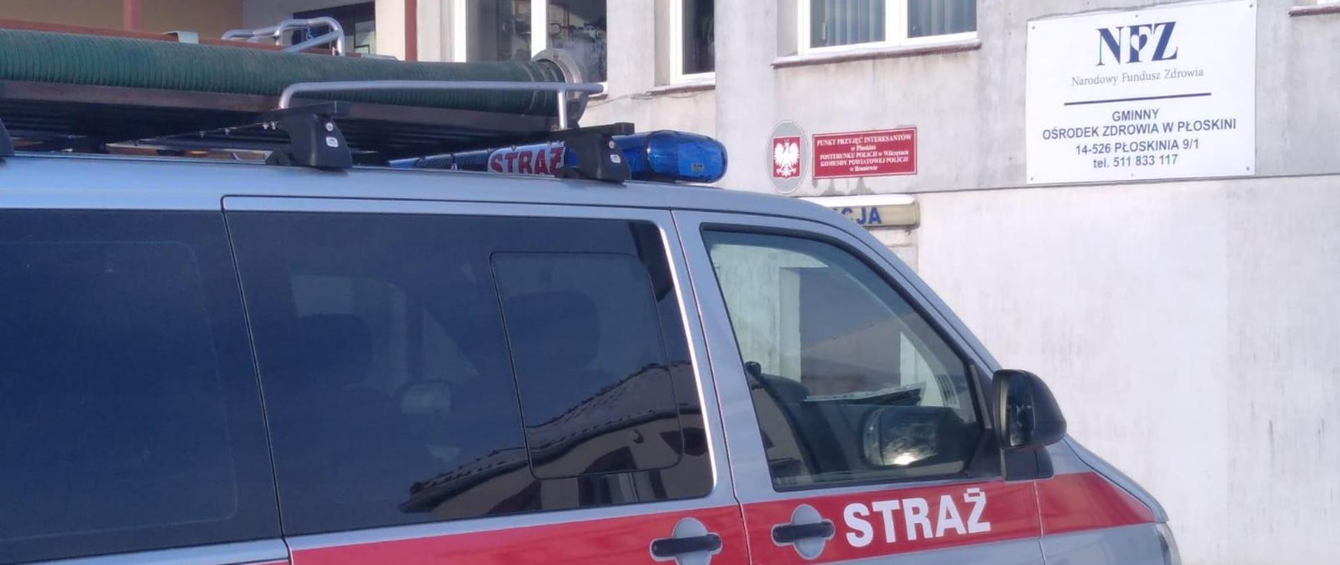 na zdjęciu budynek gminnego ośrodka zdrowia a przy budynku samochód pożarniczy przodem do budynku. Samochód koloru szarego z czerwonym pasem na całej długości z białym napisem STRAŻ. Na drzwiach pojazdu herb gminy Płoskinia i numery operacyjne jednostki OSP Dąbrowa. 