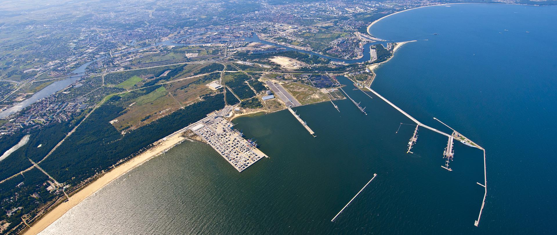 Na zdjęciu widać nabrzeże portowe w Gdańsku. Po prawej stronie można zauważyć Zatokę Gdańską, po lewej plażę wraz z portem. Pogoda jest słoneczna.