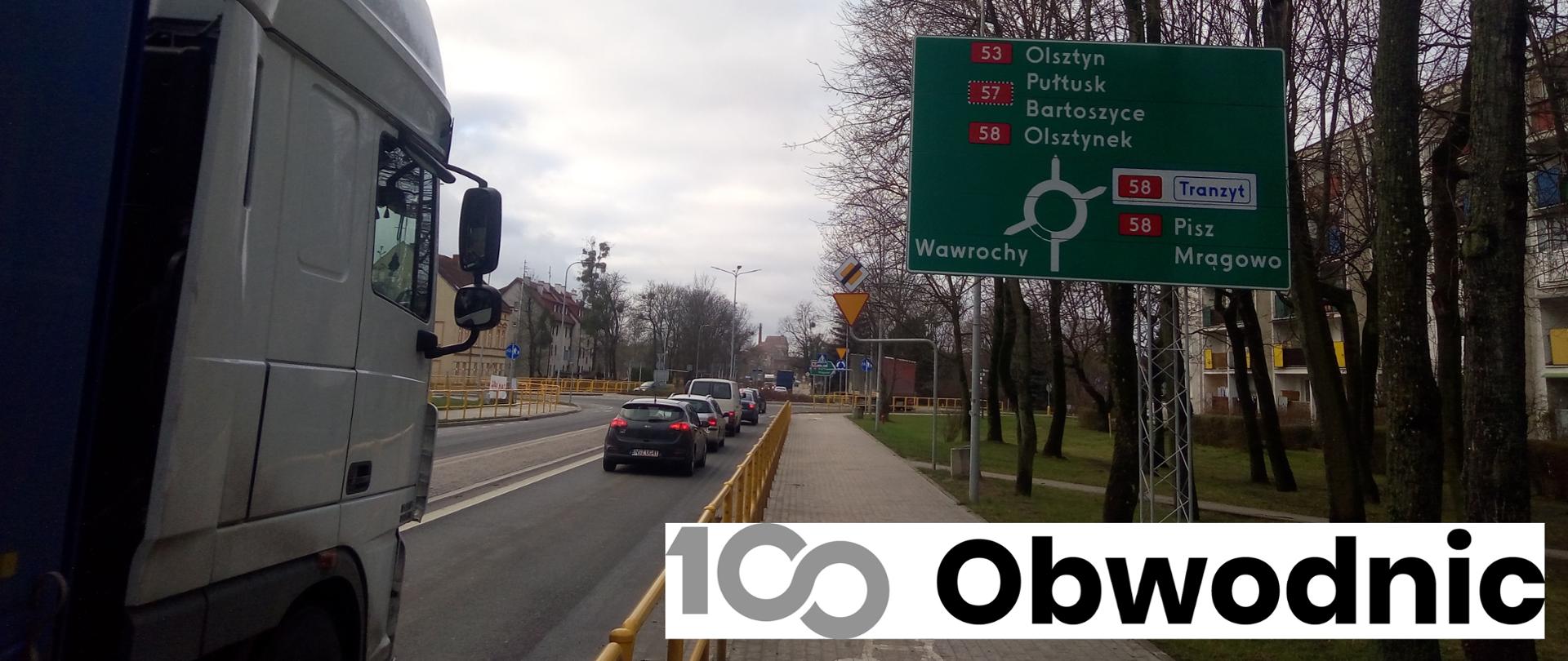 Ulica w centrum miasta. jedzie nią sznur samochodów. Na pierwszym planie ciężarówka oraz tablica drogowskazowa.