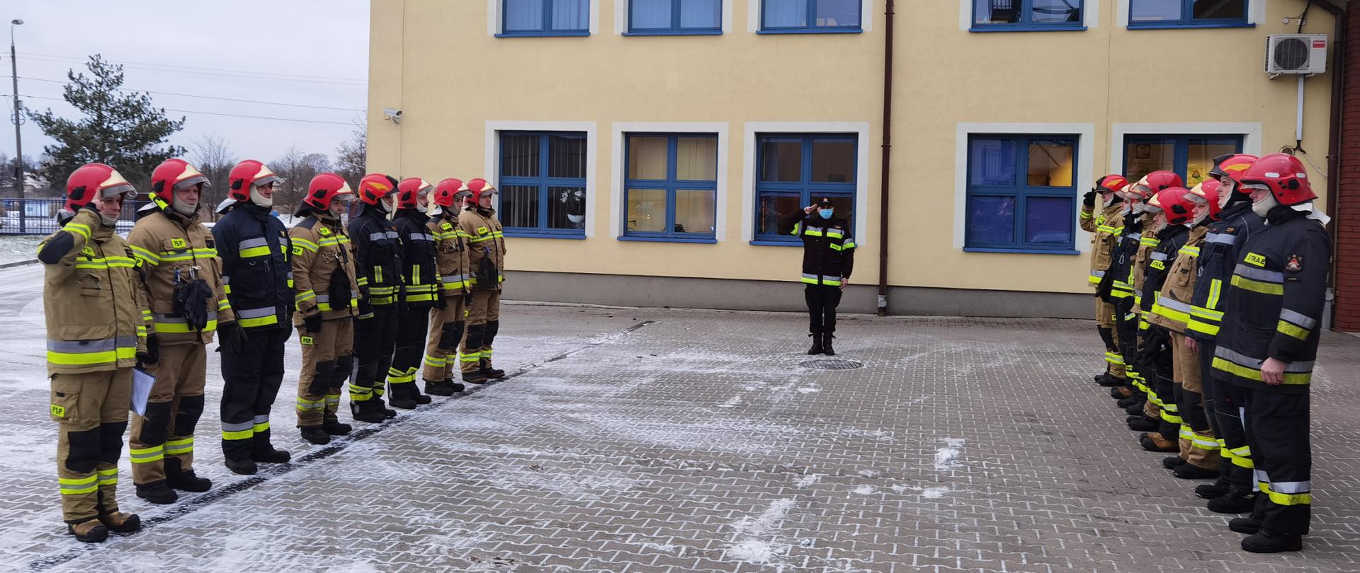 W dwóch kolumnach stoją strażacy żegnający dw Jednostki Ratowniczko Gaśniczej. Po środku stoi dw JRG