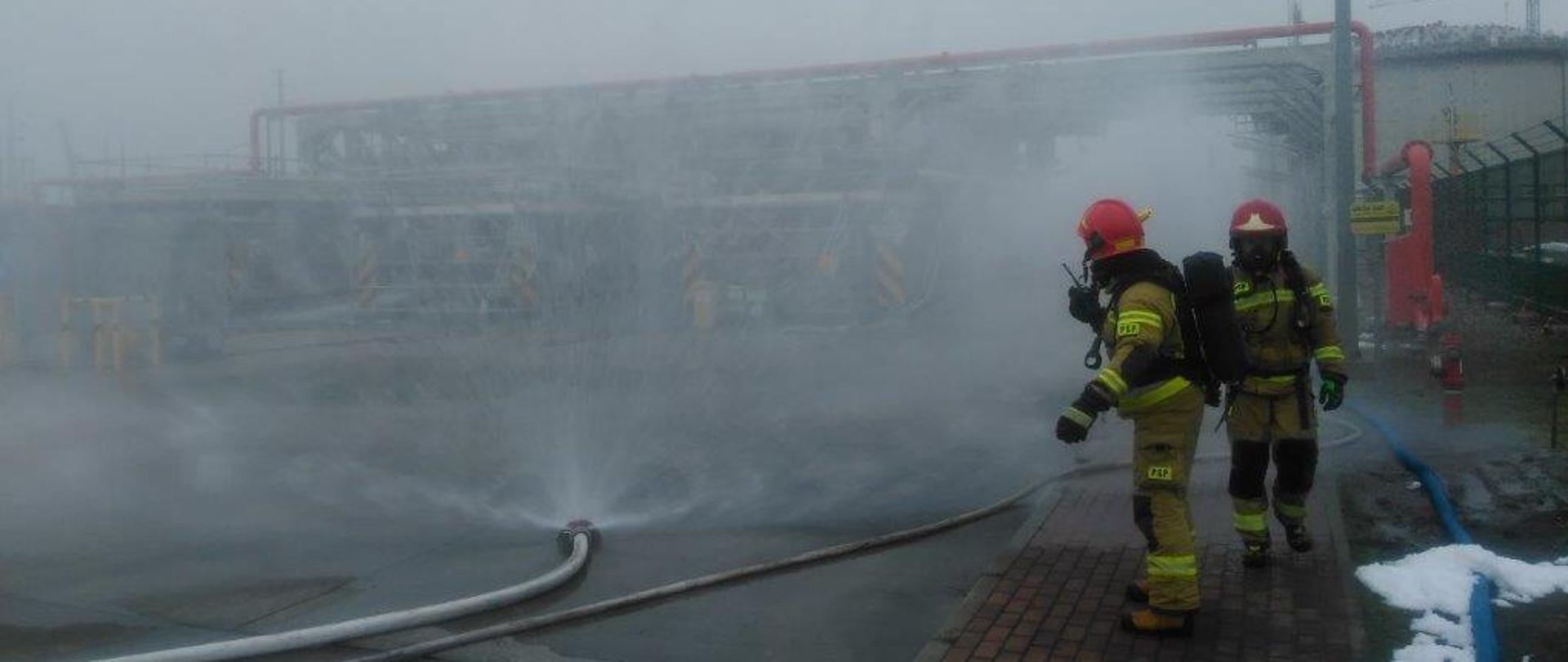Zdjęcie przedstawia dwóch strażaków w aparatach ochrony dróg oddechowych, w piaskowych ubraniach specjalnych oraz czerwonych hełmach. W tle widać kurtyny wodne podłączone do lini wężowych, z których wylatuje woda tworząc tzw. kurtynę wodną. Na dalszym planie instalacja Terminalu LNG.