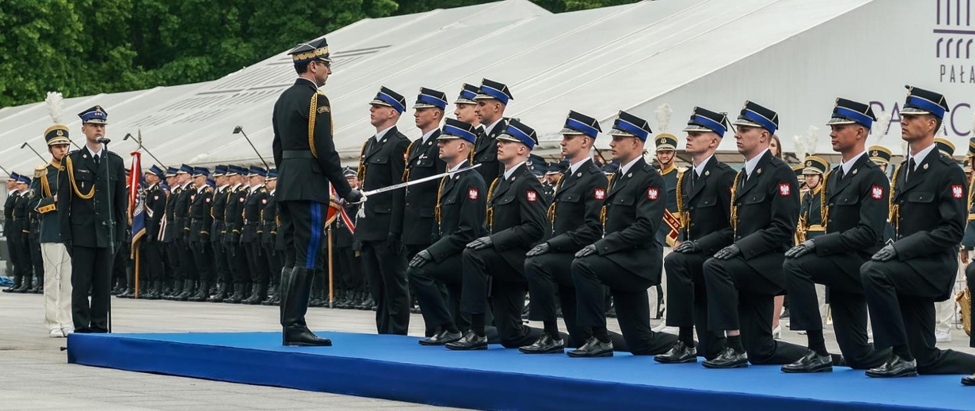 Oficer w galowym mundurze pasuje szablą klęczących podchorążych w czarnych mundurach z biało-czerwonymi flagami na rękawach podczas uroczystej ceremonii wojskowej. W tle stoi szyk żołnierzy oraz orkiestra wojskowa.