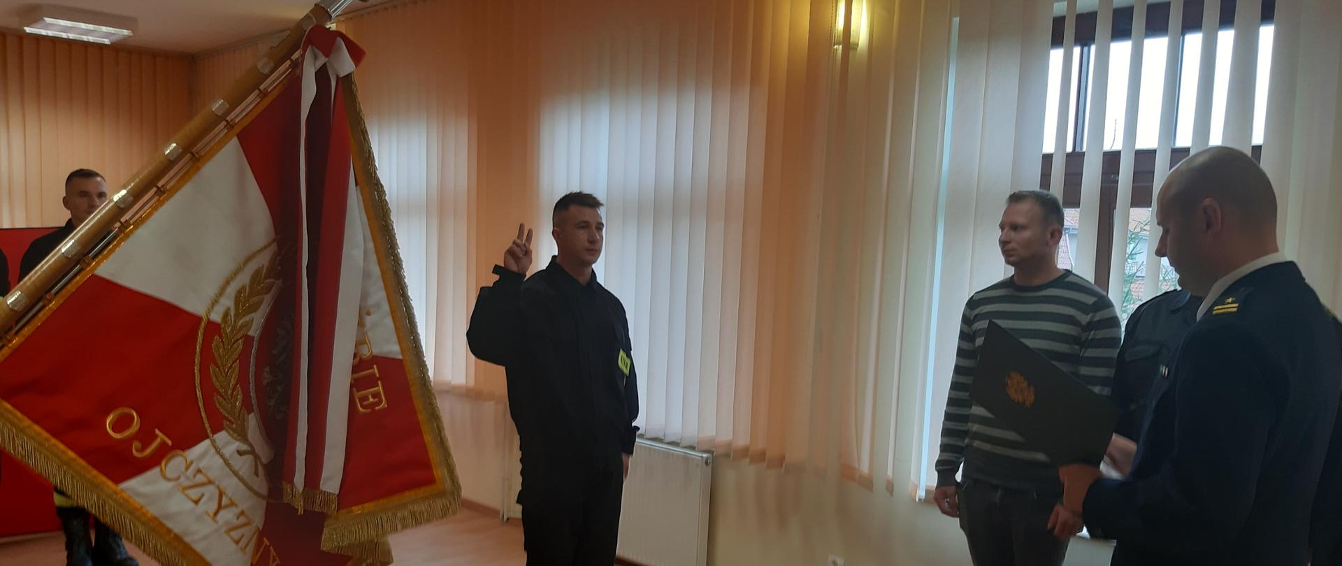 Na świetlicy strażak w ubraniu koszarowym stoi obok sztandaru i mówi rotę ślubowania. Obok niego widoczni są również inni strażacy i pracownik cywilny. 
