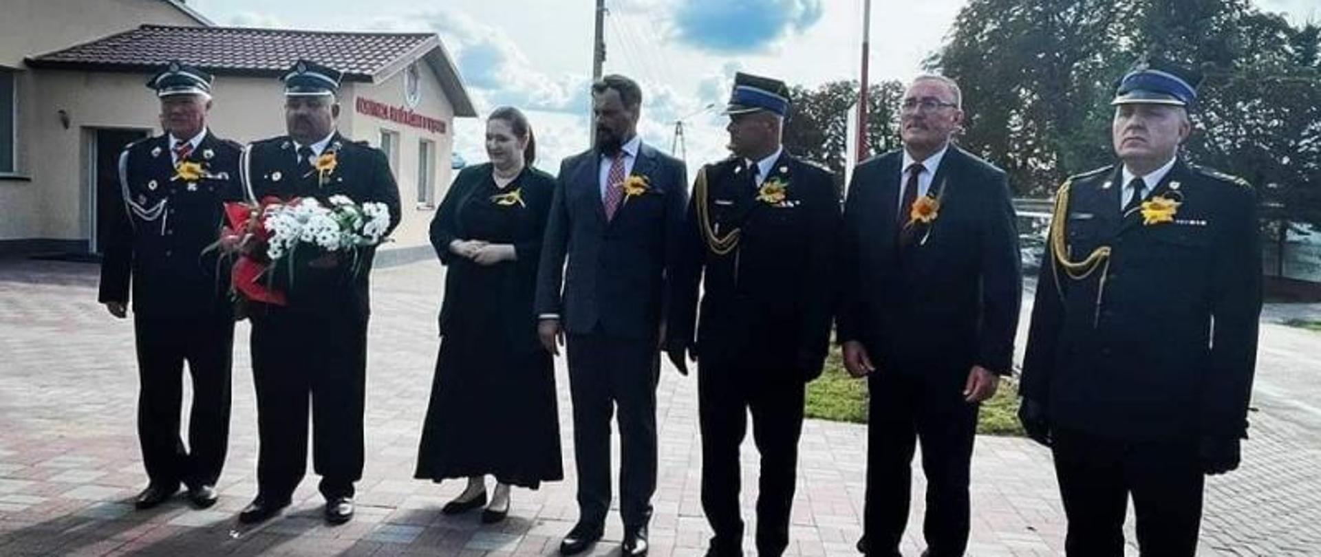 Na zdjęciu, w luźnym szeregu stoją strażacy w umundurowaniu galowym, w towarzystwie Pani minister. Wszyscy mają na piersiach kotylion dożynkowy.