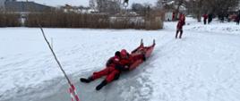 Po lodzie, na zamarzniętym zbiorniku wodnym, strażacy ciągną "sanie lodowe", na których znajduje się dwóch strażaków. Strażacy ubrani są w skafandry wodoszczelne. Na brzegu zbiornika znajdują się osoby obserwujące manewry. W oddali widoczne są pojazdy strażacki i zabudowania