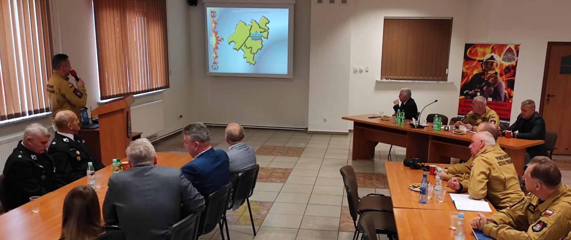 Komendant Powiatowy PSP Skarżysko-Kamienna przestawia naradę roczną za rok 2023r. Wszyscy siedzą przy stołach na świetlicy KP PSP Skarżysko.